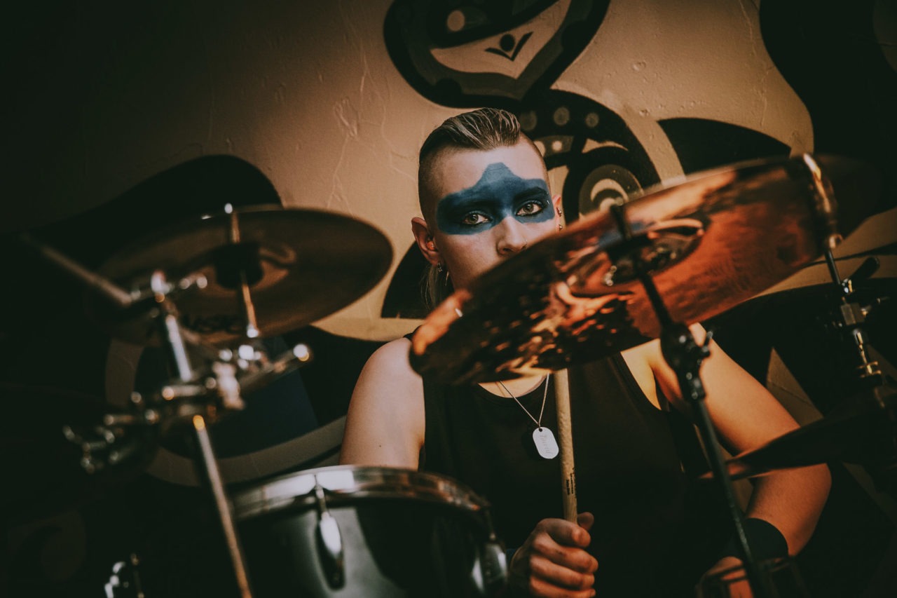 Female drummer with blue face paint staring into the camera from behind kit