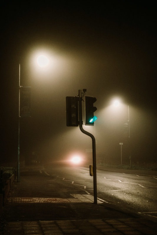 A traffic light on green stood on the side of a foggy road with street lamps emitting hazy glow