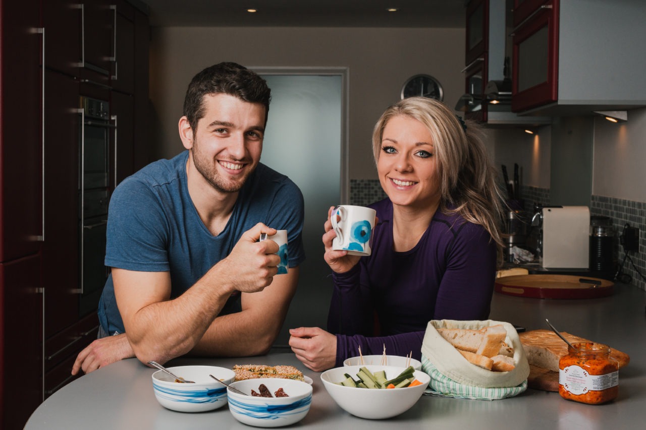 Couple stood in kitchen smiling with delicious looking food in the foreground