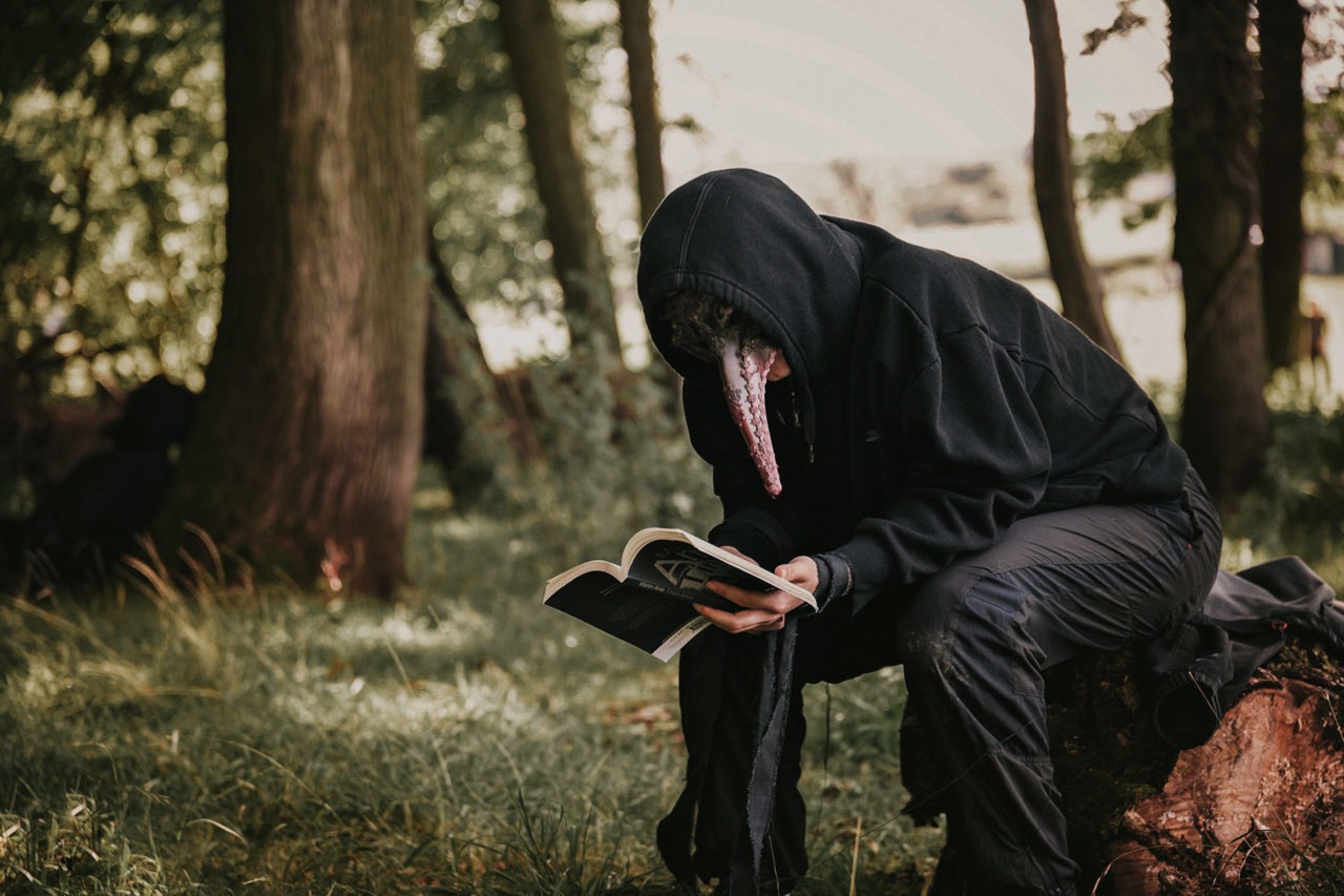 Creepy hooded character with masquerade mask sitting on tree stump reading a book