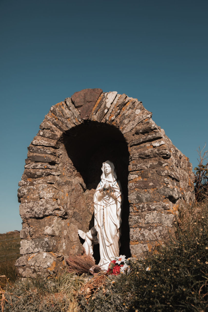 Statur of St Non is small stone archway against a blue sky