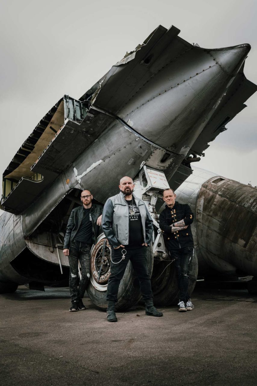 Trio of musicians stood in front of an old jet engine on a bleak day