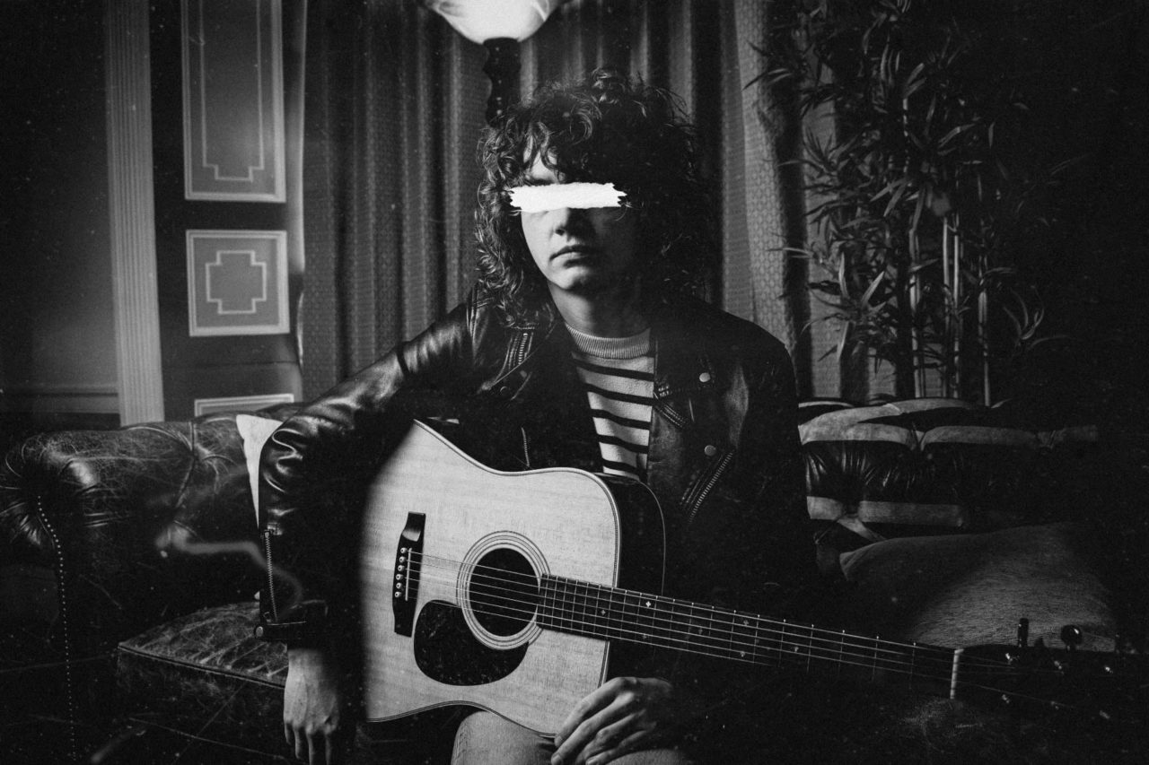 Musician in black and white sat with guitar with scratched out eyes