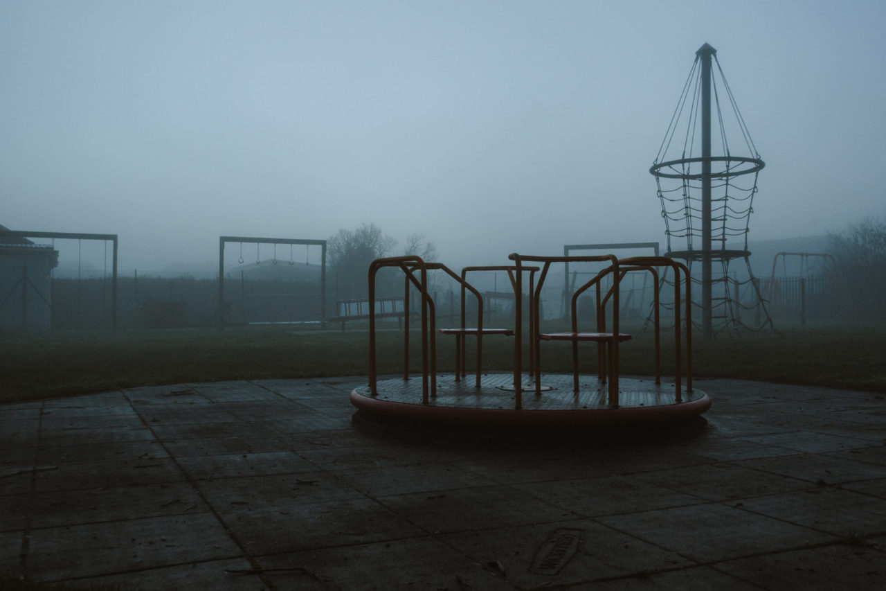 An empty child's park on a cold, foggy day, with an empty roundabout in the foreground
