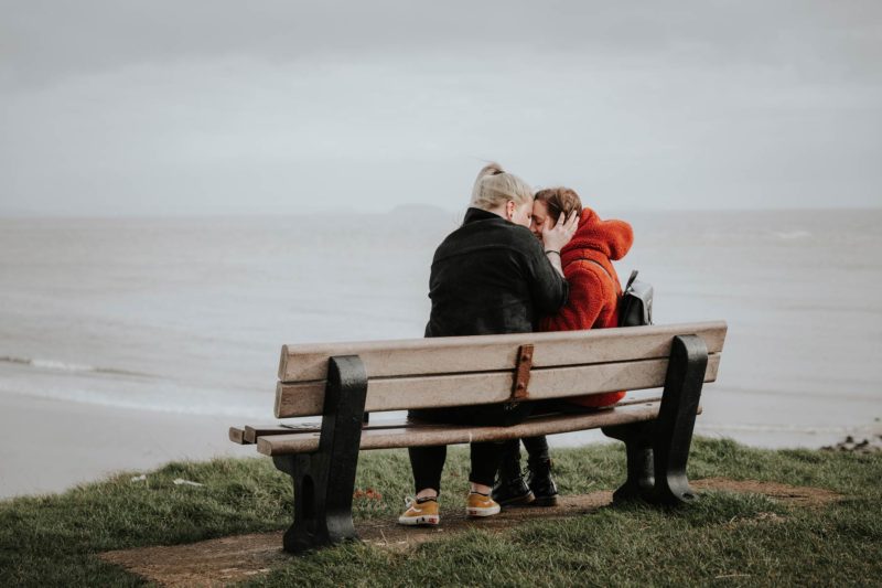 Couple sat happily on a bench with the sea behind them