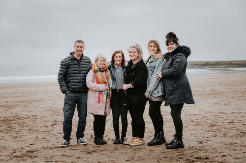 Newly engaged couple stood on beach with happy family