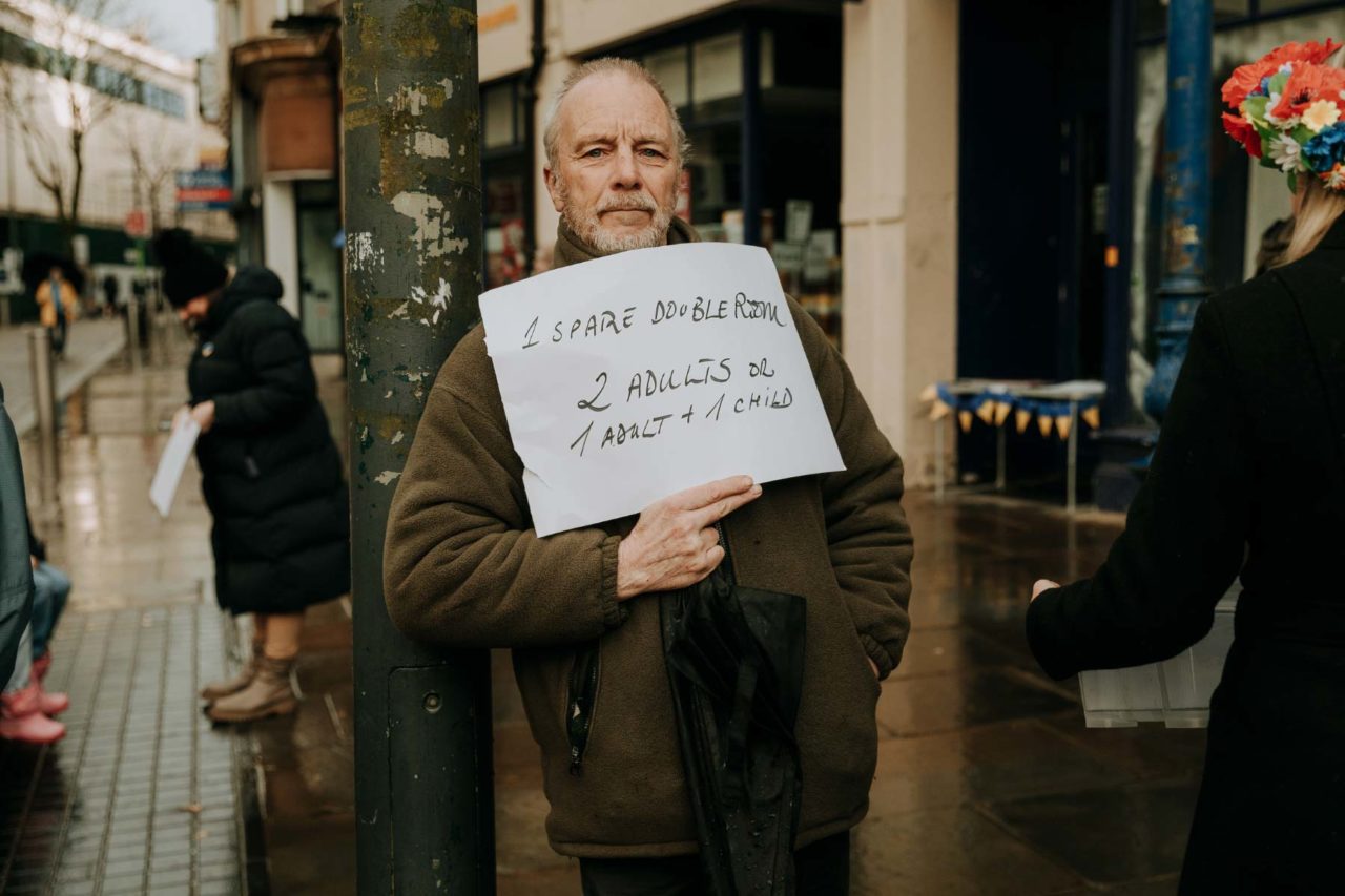 A middle aged man holding up a sign offering accomodation to refugees