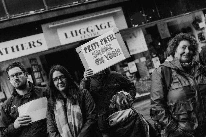 A man obscures his face with a sign reading "Shame on you Priti Patel"