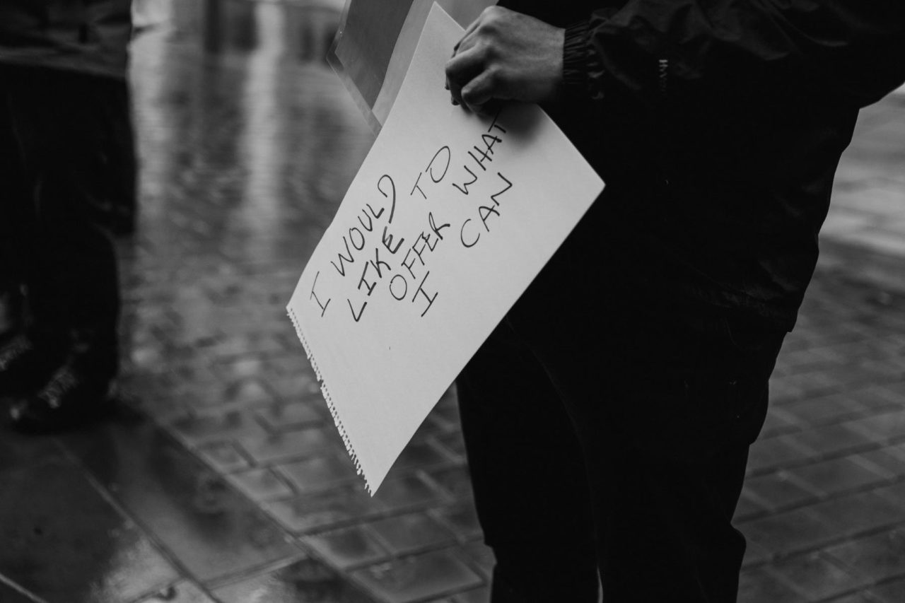 Black and white close up of a sign reading "I want to help in any way I can"