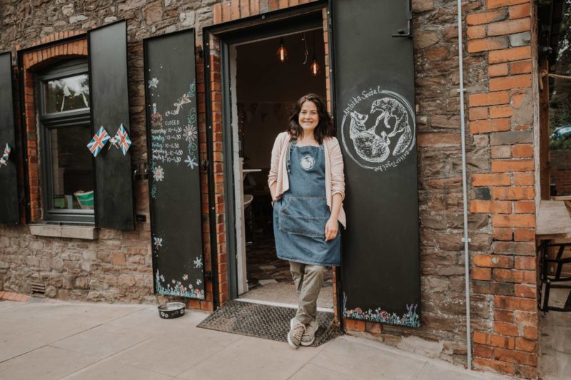 Female small business owner leant against the door of her café smiling. Shot for Ambika Social coffee shop.