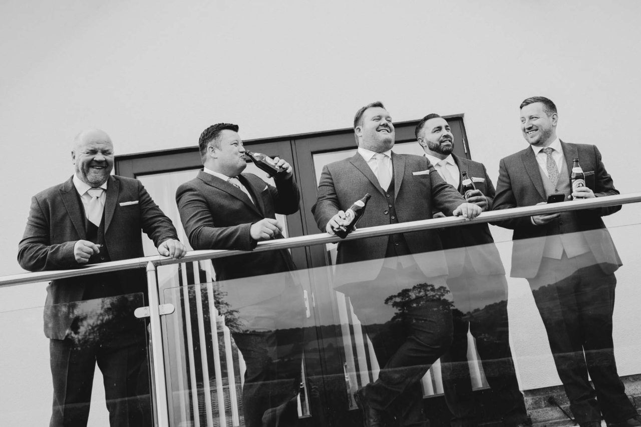 A group of best men stood drinking on a balcony laughing and enjoying themselves