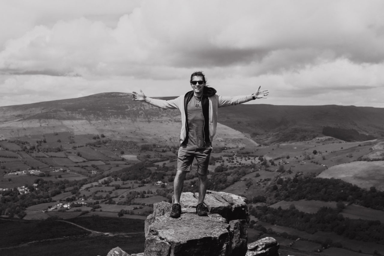 A black and white portrait of Tom Damsell stood on top of a mountain with the Brecon Beacons landscape in the background
