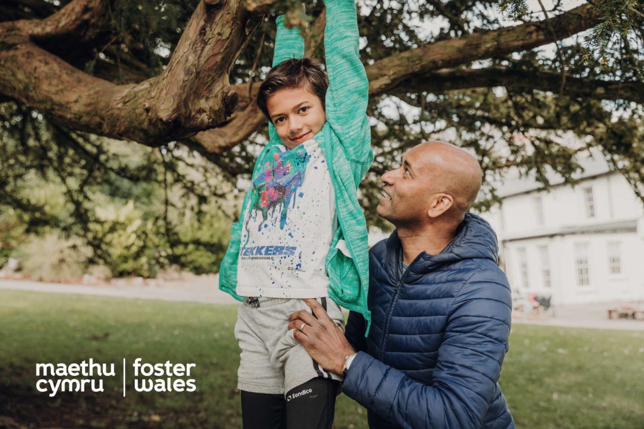 Happy looking boy hangs from a tree branch while his foster father supports his weight