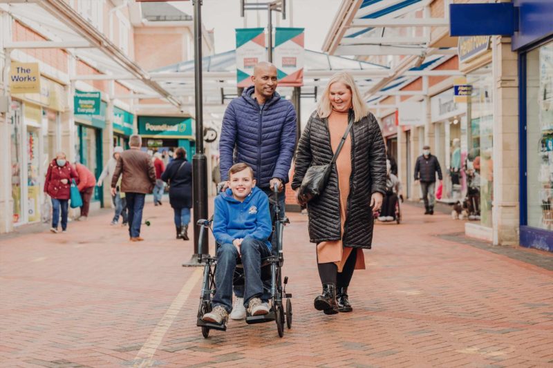 A mixed race couple push their foster son's wheelchair through a busy shopping precinct