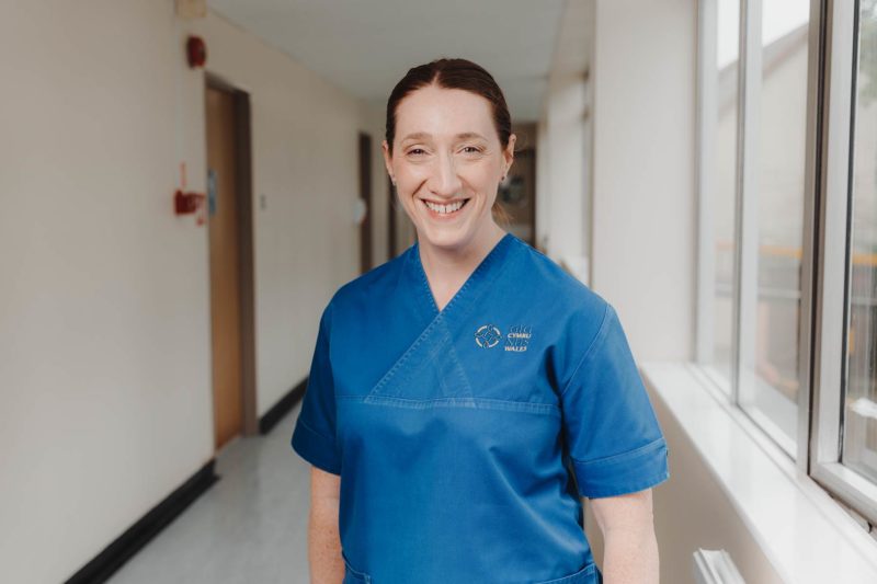 NHS nurse stood in a blue scrubs smiling in a hospital corridor