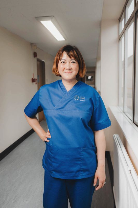 NHS nurse stood in blue scrubs smiling in a hospital corridor