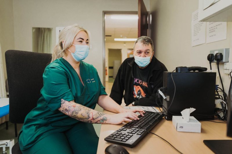 NHS nurse in green scrubs sat smiling with a male patient