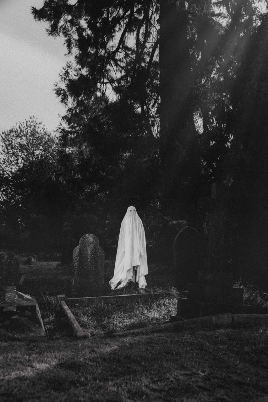 Figure draped in a white sheet, stood in a graveyard at early morning in black and white