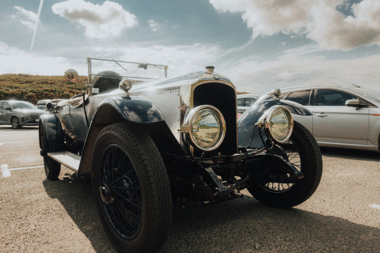An open top classic car sat under a blue sky with light glinting off the paint work