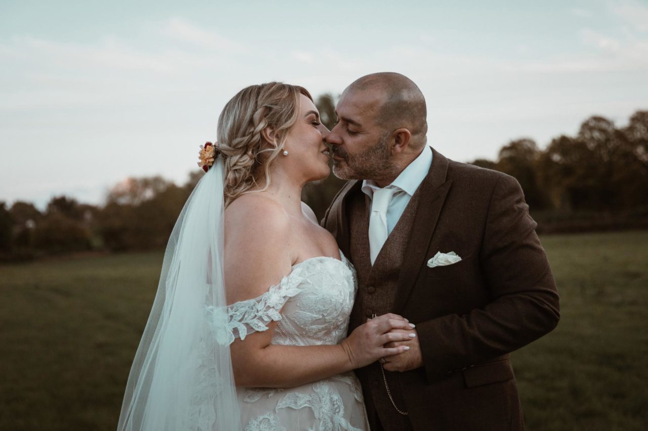 Newly weds stood in a field at dusk sharing a kiss