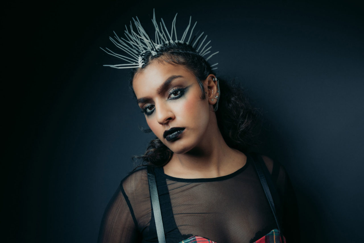 Model in alternative makeup and punk style hairstyle stood against a dark background posing in studio shoot