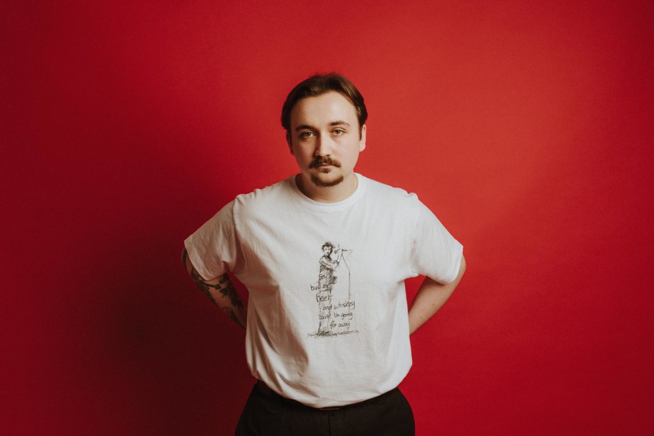 Man in a white t-shirt stood with hands behind his back against a red backdrop