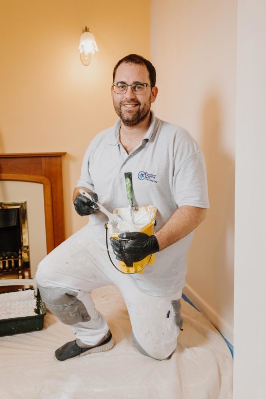Decorator knelt smiling at camera while holding a yellow paint tub and brush