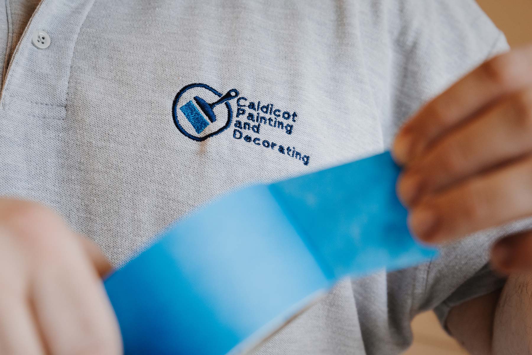 Close up of shirt with company logo on a shirt wih blurry hands holding blue tape in the foreground