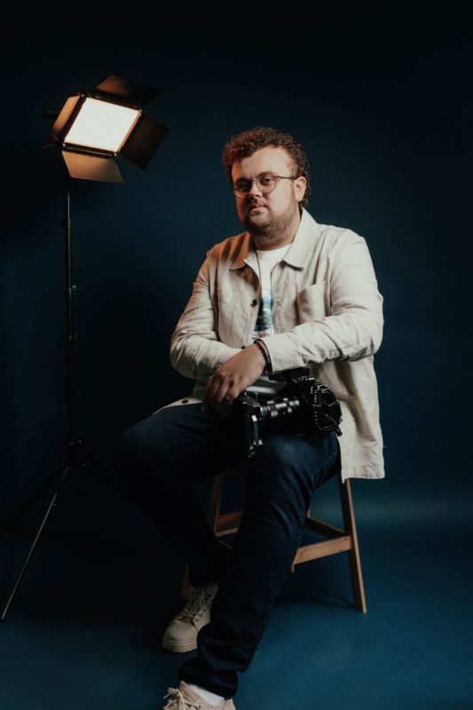 Will Langley of CBloc Productions sat for a studio shoot holding a camera, lit with a prop light