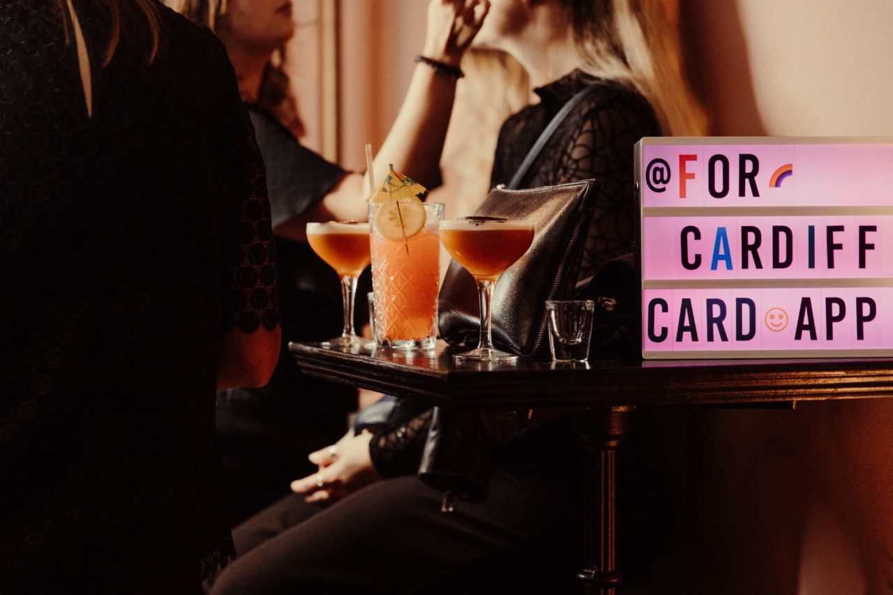 Close up of cocktail glasses sat on a table next to a FORCardiff sign with people in the background