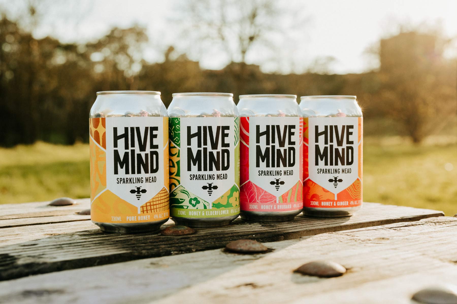 4 colourful cans of Hive Mind sparkling mead sat on a bench at sunset with a blurry country park in the background