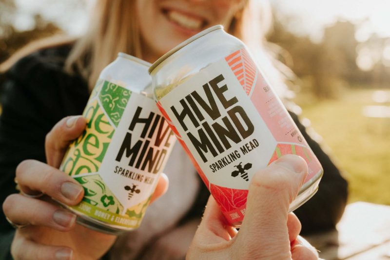 Close up of two colourful cans of Hive Mind sparkling mead with a smiley girl in the background, taken at sunset
