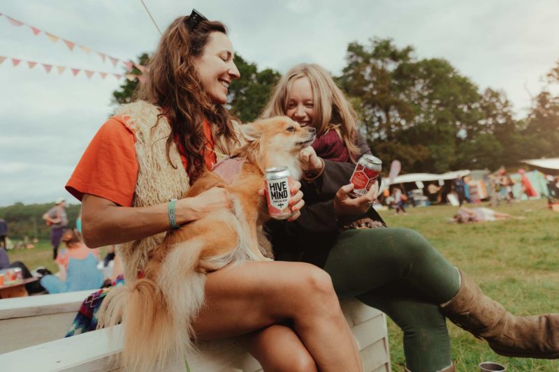 Two woman sat on a bench with a dog at Green Gathering Festival having fun and holding cans of Hive Mind mead