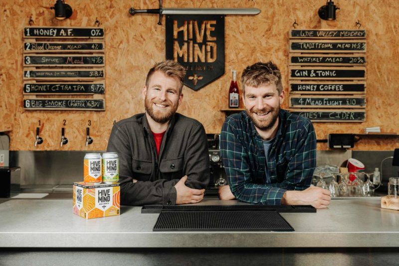 Wye Valley Meadery owners; Kit and Matt, stood smiling behind the bar with Hive Mind cans sat next to them and brand signage behind.