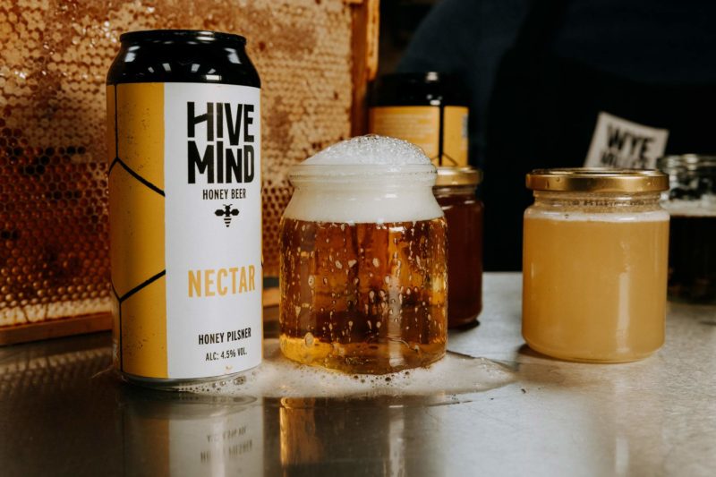 Product shot of a jar of beer that's overflowed onto the bar stood next to a beehive and a yellow can of Hive Mind Nectar