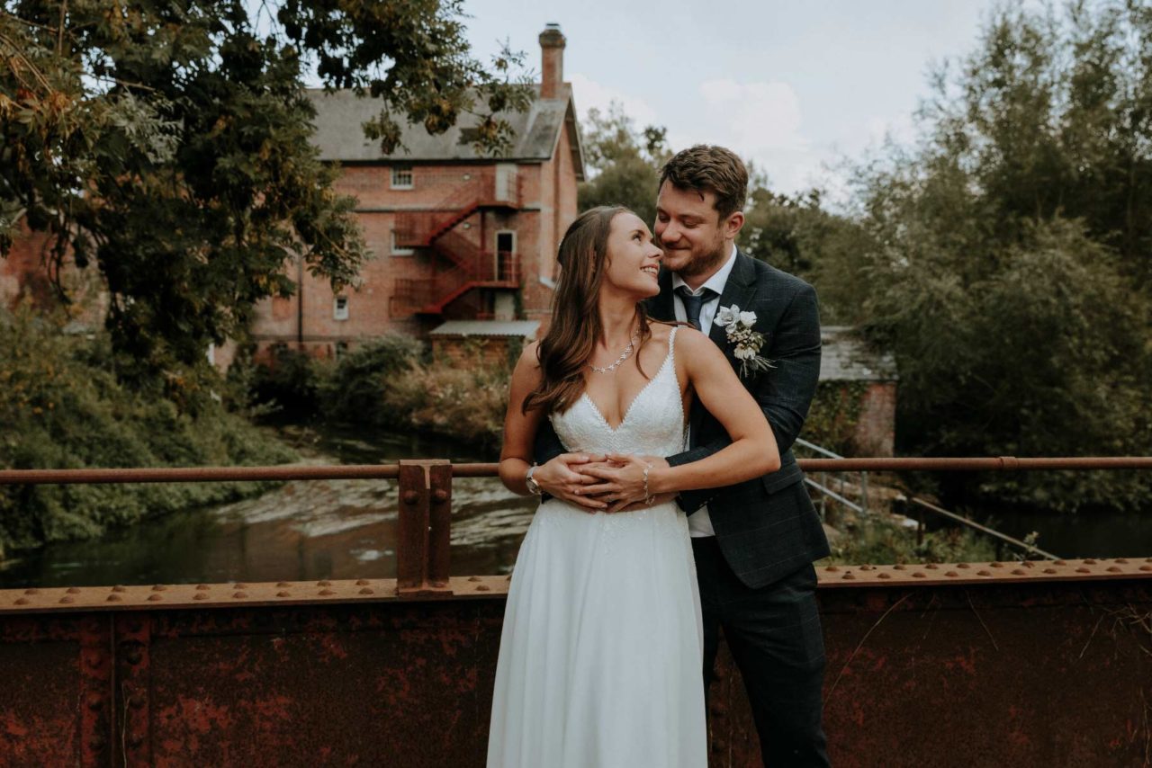 Newly weds stood on an iron bridge with beautiful old mill in the background