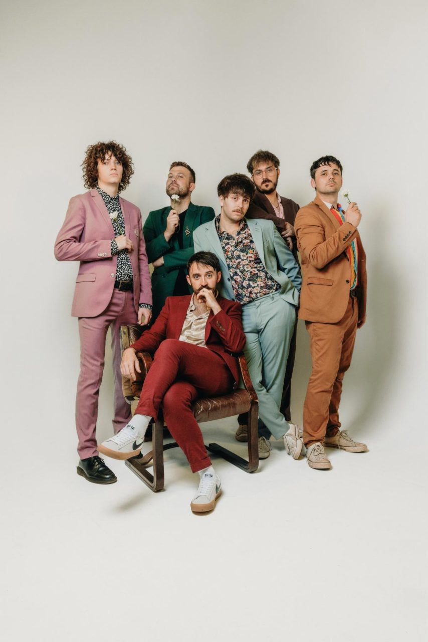 Rusty Shackle stood against a white backdrop in coloured suits posing with white flowers