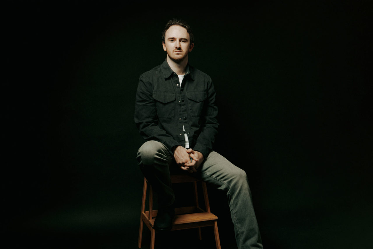 A young man sat on a stool in front of a black studio backdrop with dramatic moody lighting and heavy shadow