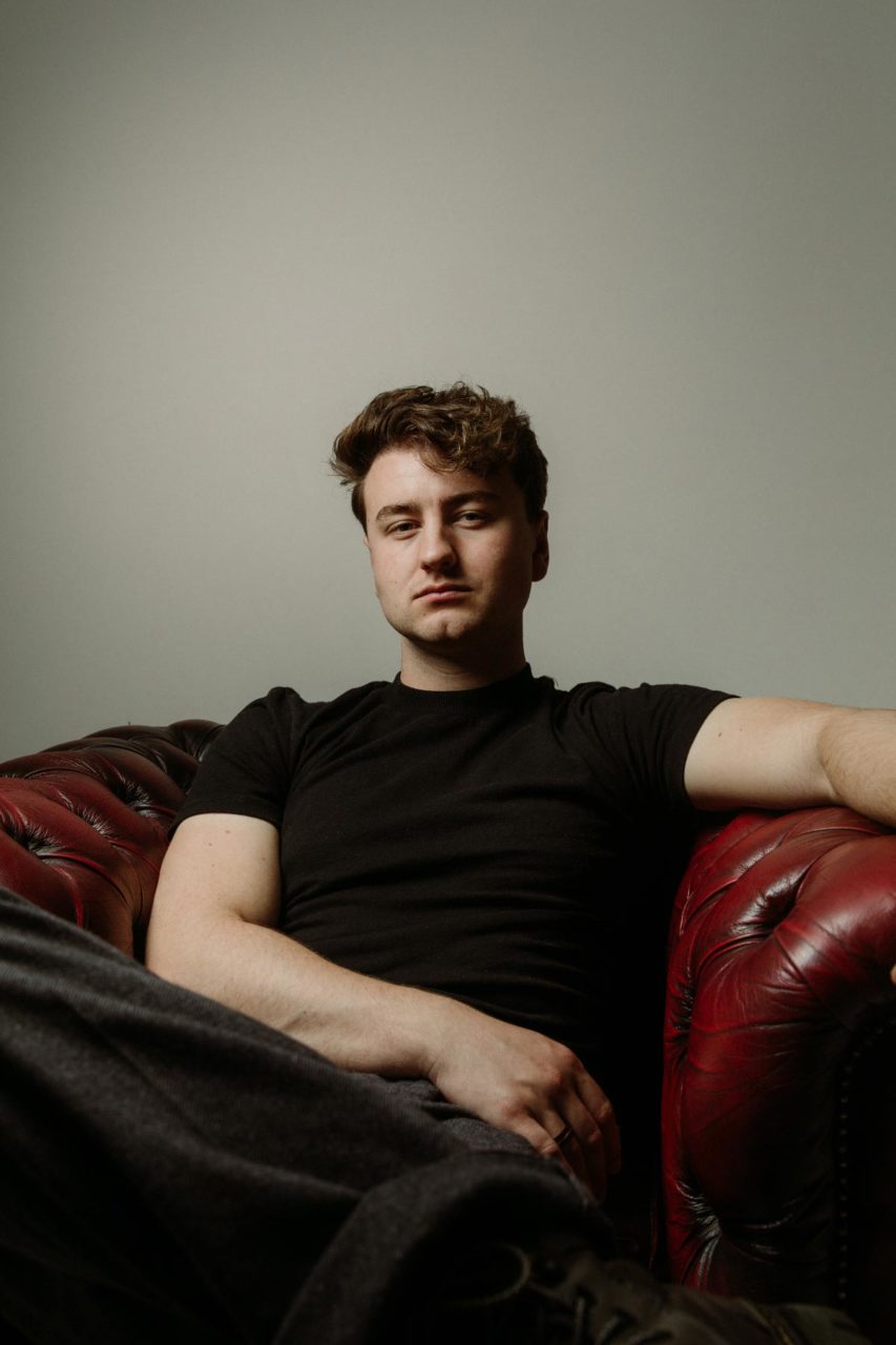 High contrast colour portrait of Tom Auton sat on an old red leather chair on set at Digichemistry studio