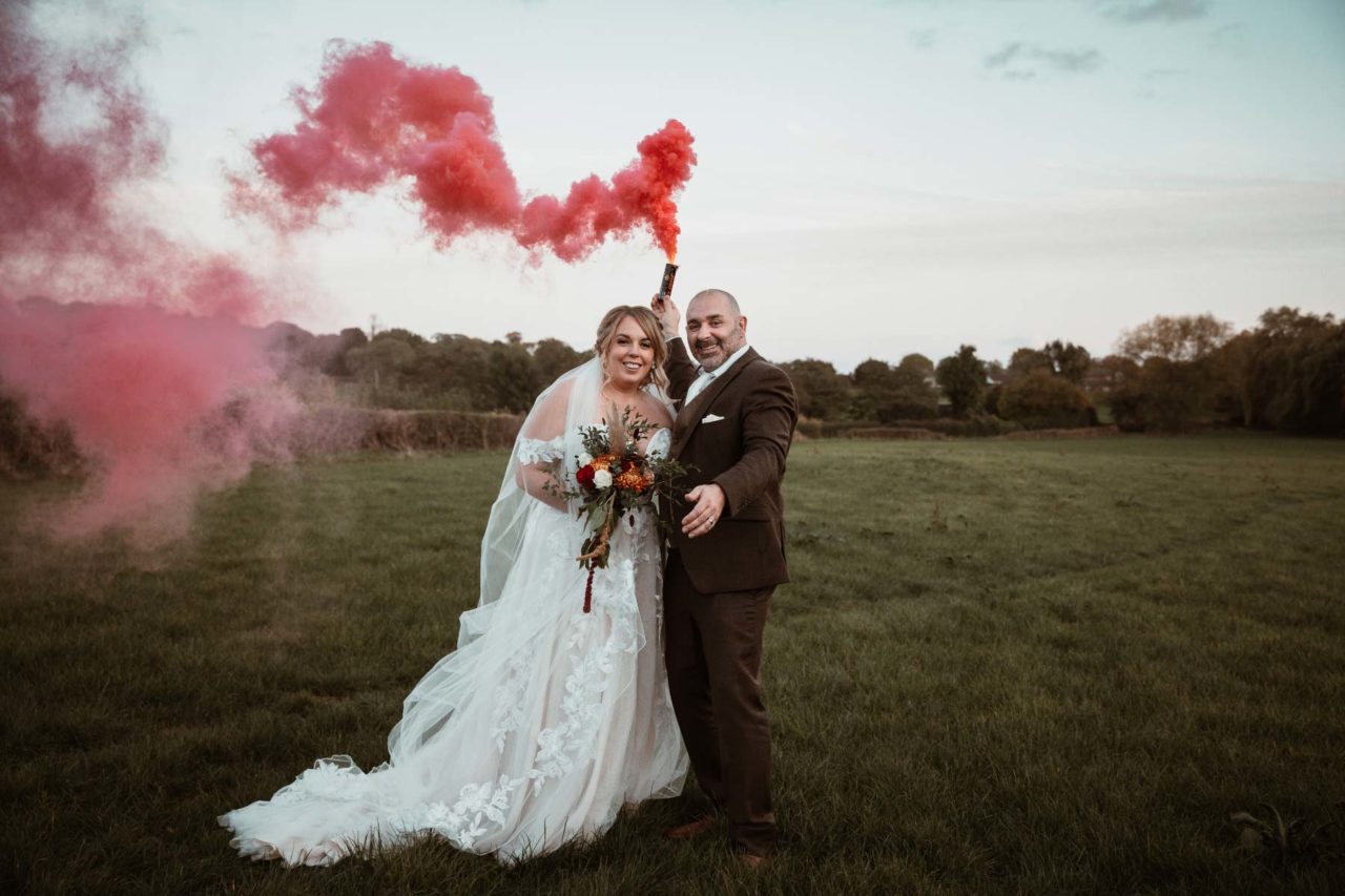Newly weds stood in a field at dusk laughing with a red smoke bomb going off behind them