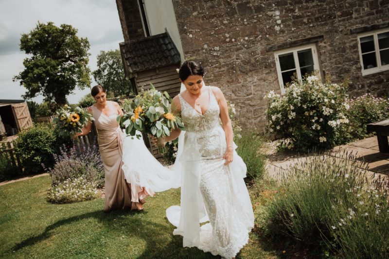 Bride walking through a garden while her bridesmaid follows behind carrying her veil