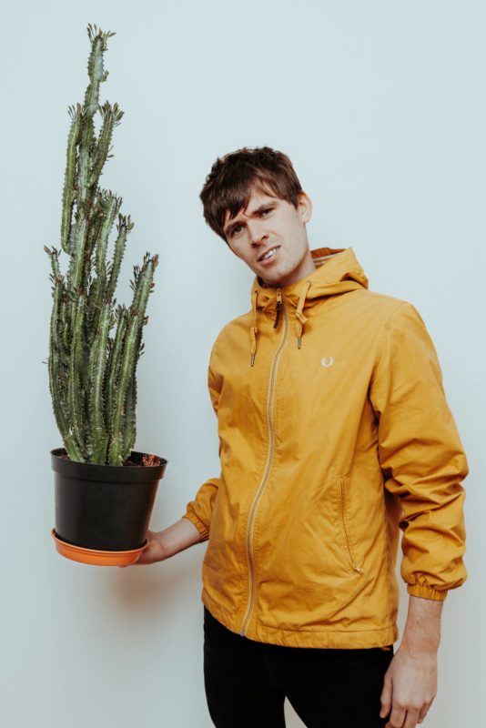 Jack Perrett stood holding a cactus wearing a bright yelllow jacket. Shot at Citrus Studio, Cardiff.