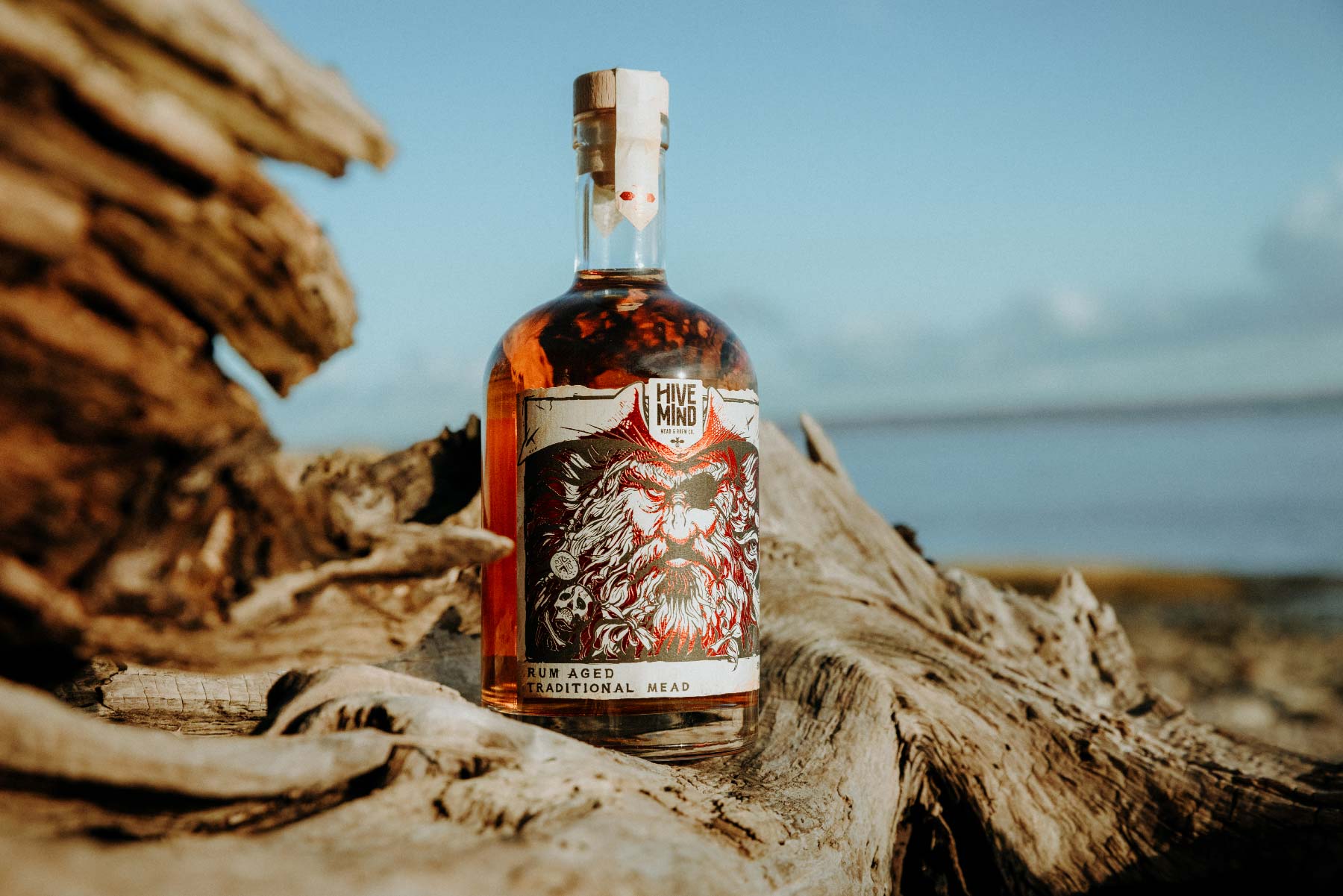 Promotional product image of a bottle of Hive Mind rum aged mead sat on a piece of drift wood, with blue skies and see in the background. Shot as part of a promotional marketing shoot, Nov 2024.