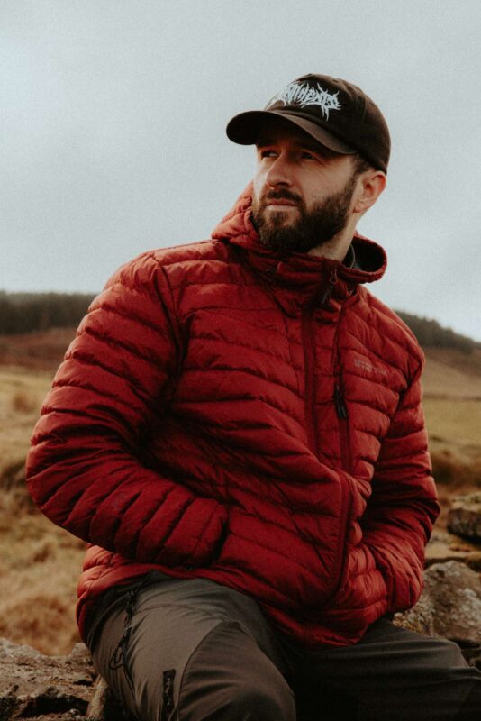 Portrait of a man wearing a red Mountain warehouse jacket, sat outdoors in the wild of Dare Valley Country Park, South Wales.